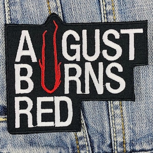 以下が含まれることがあります： 白で刺繍された「AUGUST BURNS RED」の文字が入った黒いパッチ。赤い様式化された「U」の形が中央にあります。パッチは長方形で、右側にユニークな形をしています。