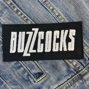 May include: A black rectangular patch with the band name "BUZZCOCKS" embroidered in white, stylized block letters. The patch is set against a blue denim background, with visible stitching and seams.