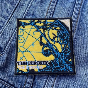 Può includere: Toppa ricamata con il nome della band "THE STROKES" in testo bianco su un bordo nero. La toppa ha uno sfondo giallo con elementi di design blu astratti. La toppa quadrata è su una giacca di jeans.