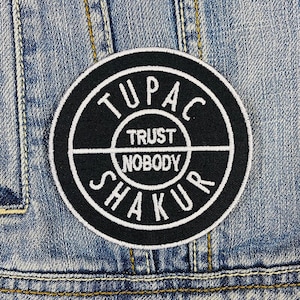 Puede incluir: Un parche bordado en blanco y negro con el texto "TUPAC TRUST NOBODY SHAKUR" en un diseño circular. El parche está cosido en una chaqueta vaquera.