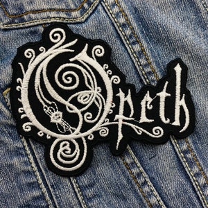 Peut inclure: Un écusson brodé noir et blanc avec le nom du groupe "Opeth" dans une police gothique. L'écusson présente un motif orné avec des motifs tourbillonnants et est posé sur une veste en jean.
