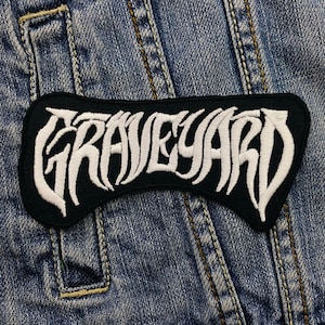 Può includere: Toppa nera con la scritta "GRAVEYARD" ricamata in bianco con un carattere gotico stilizzato. La toppa è cucita su una giacca di jeans, mostrando la sua applicazione su abbigliamento o accessori.