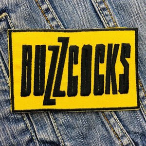 May include: A rectangular yellow patch with the embroidered text "BUZZCOCKS" in bold black letters. The patch is set against a denim background, showcasing the contrasting colors and textures of the patch and the fabric.