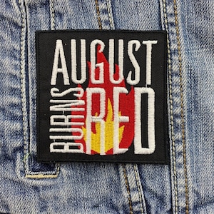 以下が含まれることがあります： 白文字で「AUGUST BURNS RED」と書かれた黒い正方形のパッチ。赤、黄、オレンジ色の炎のデザインが施されています。パッチはデニムジャケットに縫い付けられています。