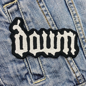 May include: A black and white embroidered patch with the word "Down" in a gothic font. The patch is rectangular with a black border and is sewn onto a denim jacket.