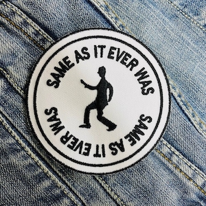 Può includere: Una toppa bianca circolare con testo nero e una silhouette nera di una persona. Il testo attorno al bordo recita "SAME AS IT EVER WAS". La toppa è su uno sfondo di denim.