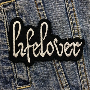 Op de afbeelding: Een zwart geborduurd embleem met het woord "lifelover" in witte cursieve letters. De patch heeft een golvende rand en is geplaatst tegen een blauwe denim achtergrond.