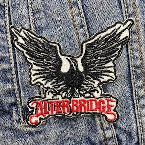 Op de afbeelding: Geborduurde patch met een zwart-witte adelaar met uitgespreide vleugels. De patch heeft de tekst "ALTER BRIDGE" in het rood, met een witte rand. De patch is bevestigd op een blauw spijkerjasje.