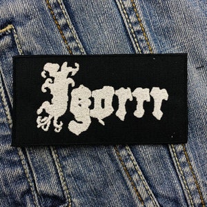 May include: A black rectangular patch with the white embroidered word "Jgorr". The patch is set against a blue denim background. The lettering has a gothic, stylized design.