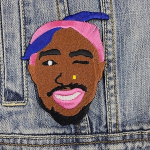 Puede incluir: Parche bordado con un retrato estilizado de una persona con piel morena, cabello rosado y una diadema azul. El parche tiene un ojo guiñando y un piercing en la nariz amarillo. El parche está cosido en una chaqueta vaquera.