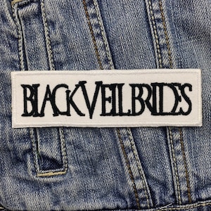 Könnte beinhalten: Ein rechteckiger weißer Aufnäher mit dem Text "BLACK VEIL BRIDES" in Schwarz gestickt, vor einer Jeansjacke. Der Text ist in einer fetten, stilisierten Schriftart gehalten.