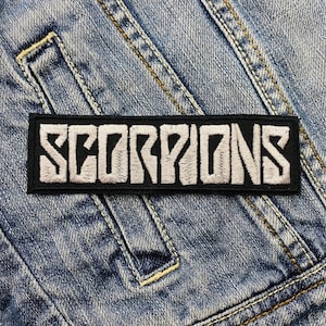May include: A rectangular black patch with the word "SCORPIONS" embroidered in white, displayed on a denim jacket. The patch features a bold, stylized font, and the denim jacket has visible stitching.