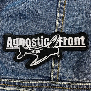Puede incluir: Parche bordado en blanco y negro con el texto "Agnostic Front" y un diseño de avión estilizado. El parche rectangular está cosido en una chaqueta vaquera. El parche mide aproximadamente 10 cm de largo.