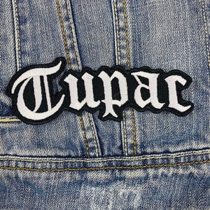 Puede incluir: Parche bordado con el nombre "Tupac" en letras góticas blancas sobre un fondo negro. El parche está cosido en una chaqueta vaquera azul, mostrando el diseño y la colocación del parche.