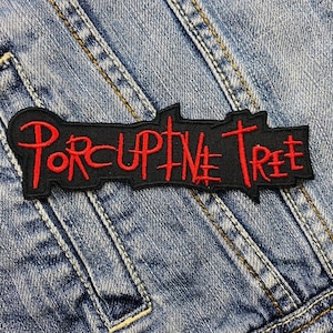 May include: Black embroidered patch with the words "Porcupine Tree" in red, stylized lettering. The patch is rectangular with a jagged top edge and is sewn onto a denim jacket.