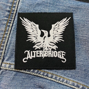 Op de afbeelding: Zwart vierkantige patch met een wit adelaarsontwerp met uitgespreide vleugels en de tekst "ALTER BRIDGE" in een gestileerd lettertype. De patch is geplaatst tegen een denim achtergrond.