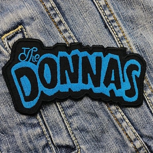 Puede incluir: Parche bordado negro y azul con el texto "The Donnas" en una fuente estilizada. El parche rectangular tiene un borde negro y está sobre un fondo de mezclilla.