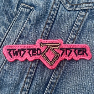 May include: A pink embroidered patch with the words "TWISTED SISTER" in black lettering. The patch features a stylized logo in the center. The patch is sewn onto a blue denim jacket.