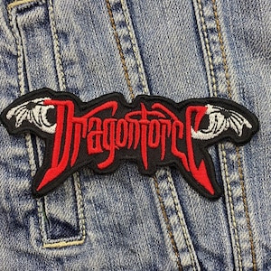 Könnte beinhalten: Ein schwarzer, gestickter Aufnäher mit dem Wort "Dragonforce" in roter, stilisierter Schrift. Der Aufnäher hat einen schwarzen Rand und ist vor einem blauen Jeanshintergrund platziert. Das Design beinhaltet flügelartige Akzente.