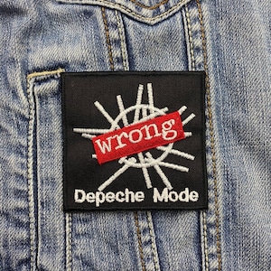 Peut inclure: Un écusson noir avec le mot "wrong" dans un rectangle rouge et les mots "Depeche Mode" en blanc. L'écusson a un motif blanc et est cousu sur une veste en jean.