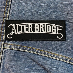 Op de afbeelding: Een rechthoekige zwarte patch met de witte tekst "ALTER BRIDGE" in een gestileerd lettertype. De patch is genaaid op een blauwe denim stof, met zichtbare stiksels en naden.