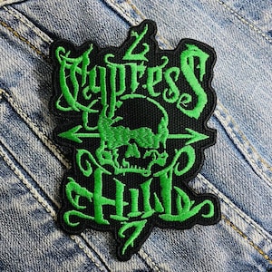 Può includere: Toppa nera con testo ricamato verde e un disegno a teschio. Il testo recita "Cypress Hill" con un teschio al centro e frecce che puntano verso l'esterno. La toppa è su uno sfondo di denim.