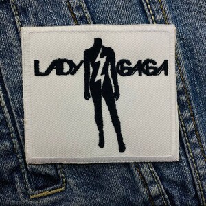 Könnte beinhalten: Ein weißer, gestickter Aufnäher mit dem Text "LADY GAGA" über einer schwarzen Silhouette einer Person mit Blitzdesign. Der Aufnäher ist rechteckig und vor einem Jeanshintergrund platziert.