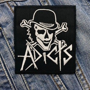 May include: Black square patch with a white skull wearing a bowler hat and crossed bones. The word "APICKS" is written in white, stylized lettering. The patch is set against a denim background.