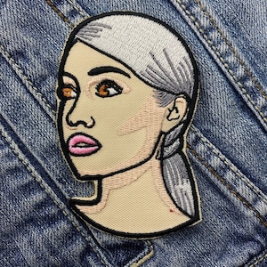 May include: Embroidered patch featuring a stylized female face with light skin, brown eyes, and pink lips. The hair is light gray, and the patch is outlined in black. The patch is set against a denim background.