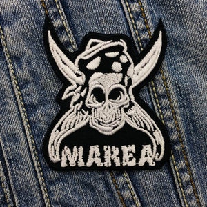 Könnte beinhalten: Schwarz-weißer, gestickter Aufnäher mit einem Totenkopf, der einen Hut und ein Bandana trägt, mit dem Wort "MAREA" darunter. Der Totenkopf hat große Augenhöhlen und ist von stilisierten Flügeln oder Hörnern umgeben. Der Aufnäher befindet sich auf einem Jeanshintergrund.