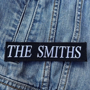 Puede incluir: Un parche rectangular negro con el texto bordado en blanco "THE SMITHS" se muestra en una chaqueta vaquera. El parche está centrado y el texto está en una fuente sans-serif. La chaqueta vaquera tiene costuras y pespuntes visibles.