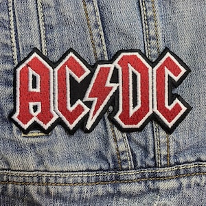 May include: Embroidered patch displaying the band name "AC/DC" in red, with a white outline and a black border. The lightning bolt in the center is also red and white. The patch is sewn onto a blue denim jacket.