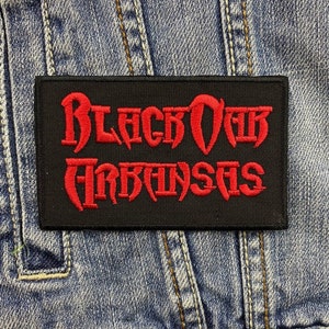 Puede incluir: Un parche rectangular negro con las palabras "Black Oak Arkansas" bordadas en rojo, con letras estilizadas. El parche está sobre un fondo de mezclilla, con costuras y bolsillos visibles.