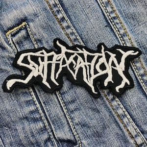 Può includere: Toppa ricamata in bianco e nero con la parola "Satan" in un carattere gotico stilizzato. La toppa è rettangolare con un bordo nero ed è cucita su una giacca di jeans.