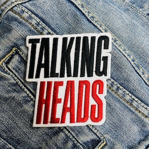 Op de afbeelding: Een rechthoekige patch met de woorden "TALKING HEADS" in zwart en rood, omlijnd met wit. De patch is genaaid op een blauwe denim stof, met zichtbare stiksels en een zakdetail.