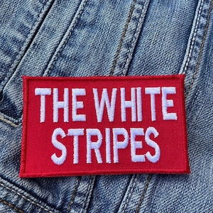 Puede incluir: Un parche rectangular rojo con texto bordado en blanco que dice "THE WHITE STRIPES". El parche se encuentra sobre un fondo de mezclilla azul, con costuras y textura de tela visibles.