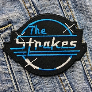 May include: A black patch featuring the words "The Strokes" in white and blue, with a distinctive design. The patch is sewn onto a denim jacket, showcasing a classic aesthetic.