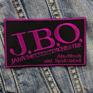 Puede incluir: Un parche rectangular negro con texto magenta bordado. El texto dice "J.B.O." en letras grandes, con "JAMES LAST ORCHESTER" y "Alschloch und Spaßdabei" debajo. El parche está sobre un fondo de mezclilla.