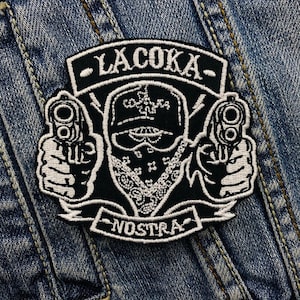 Könnte beinhalten: Schwarz-weißer, gestickter Aufnäher mit einer maskierten Figur, die zwei Pistolen hält. Der Aufnäher trägt den Text "LACOKA" über der Figur und "NOSTRA" darunter. Das Design befindet sich auf einem Jeans-Hintergrund.