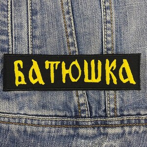 May include: A black rectangular patch with the word "БАТЮШКА" embroidered in yellow. The patch is set against a blue denim background, with visible stitching and seams.