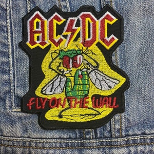 May include: Embroidered patch featuring the AC/DC logo in yellow and red, with a cartoon fly illustration. The text "FLY ON THE WALL" is below the fly. The patch is on a denim jacket.