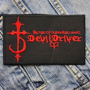 Puede incluir: Parche rectangular negro con texto bordado en rojo y un diseño de cruz estilizada. El texto dice "THE FURY OF OUR MAKERS HAND" encima de "DevilDriver" en una fuente gótica. Un pequeño símbolo está debajo.