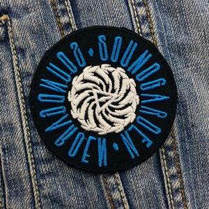 Op de afbeelding: Een ronde, zwarte geborduurde patch met de woorden "SOUND GARDEN" in blauw, die een wit, wervelend ontwerp omcirkelen. De patch is bevestigd op een spijkerjasje, wat een klassieke, vintage uitstraling geeft.