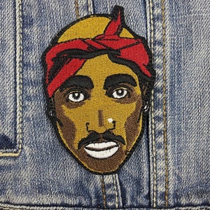 Peut inclure: Écusson brodé représentant un homme avec un bandana rouge, une moustache et une barbiche. L'écusson est cousu sur une veste en jean. Le portrait a la peau brune, des sourcils noirs et des yeux blancs.