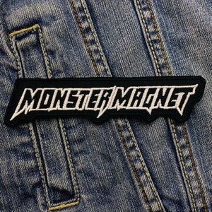 Könnte beinhalten: Ein schwarzer Aufnäher mit dem weißen, gestickten Schriftzug "MONSTER MAGNET" in einer auffälligen, stilisierten Schriftart. Der Aufnäher ist auf einer blauen Jeansjacke angebracht, mit sichtbaren Nähten und Taschen.