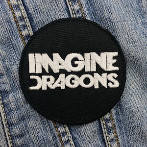 Op de afbeelding: Een ronde, zwarte patch met de geborduurde tekst "IMAGINE DRAGONS" in het wit. De patch is geplaatst tegen een blauwe denim achtergrond, wat suggereert dat deze bedoeld is om op kleding of accessoires te worden genaaid.