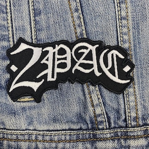 Puede incluir: Parche bordado en blanco y negro con el texto "2PAC" en fuente gótica. El parche es rectangular con un borde decorativo y está colocado sobre una chaqueta vaquera.