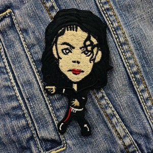 May include: Embroidered patch depicting a cartoon pop icon in a black outfit with a red stripe. The patch has black hair, a tan face, and red lips. The patch is set against a blue denim background.