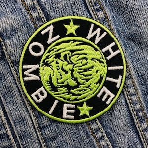 Op de afbeelding: Een ronde, zwarte geborduurde patch met de woorden "ZOMBIE WHITE" in witte letters. Het midden heeft een neon groen ontwerp. Twee groene sterren staan ​​boven en onder. De patch staat op een spijkerjas.
