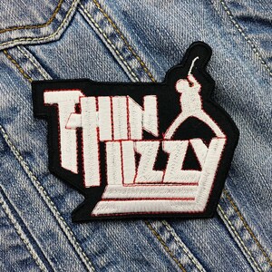 May include: Embroidered patch with the words "Thin Lizzy" in white, outlined in red, on a black background. A white silhouette of a guitarist is also featured. The patch is on a denim jacket.